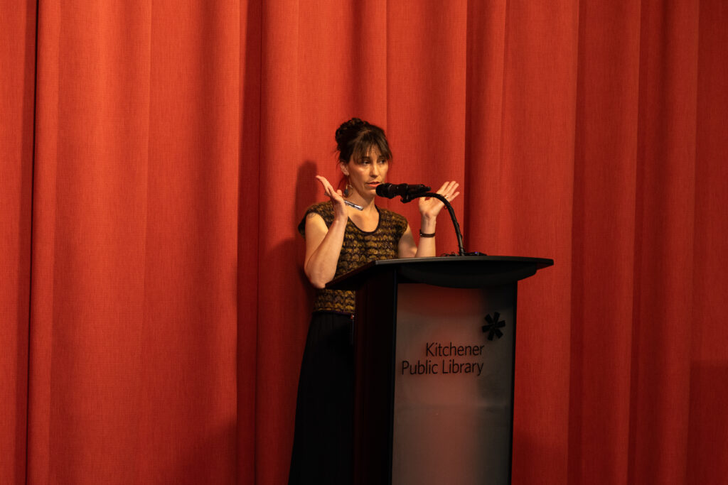Stephanie Earp standing behind a podium, talking animatedly about knitting.